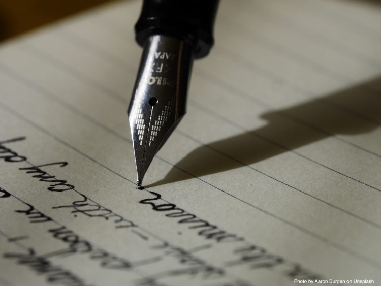 Image of an ink pen poised over a paper with cursive writing. It symbolises creativity in writing.